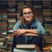 A nerdy woman sitting on the floor leaning up against a massive stack of books. She has a look of satisfaction on her face.