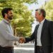 Two men dressed business casual, shaking hands on a neighborhood street.
