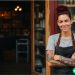 a quirky looking woman with dark red hair and tattoos standing outside of her bakery with her arms crossed, smiling at the camera.