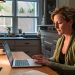 A woman in her mid 40s sitting at the dining room table with her laptop.