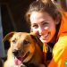 A woman wearing an orange jacket kneeling down next to a dog. Both are smiling at the camera.