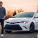 A man in his early thirties standing next to his white Toyota Camry.