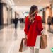A woman in a red coat walking away from the camera in a shopping mall, holding several bags of merchandise.