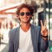 A man in his late twenties wearing a blazer and sunglasses holding up a peace sign with his fingers.