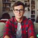 A 25 year old man in a flannel shirt and glasses that is sitting on his couch and looking at the camera. He is ready to start investing.