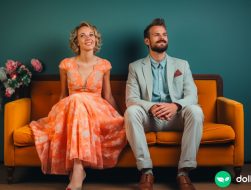 A classy married couple sitting on a couch but slightly facing away from each other. They have playful looks on their faces.