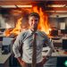 A fictitious picture of a man in a tie standing in front of cubicles that are on fire. He has a smirk on his face.