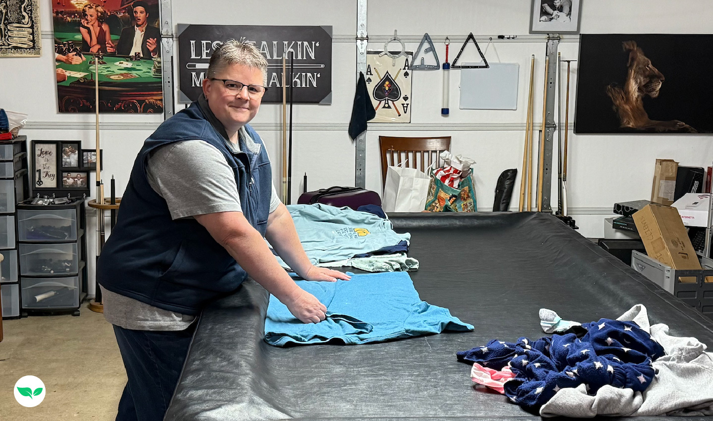 Melia Long folding laundry on a pool table in her garage, surrounded by clothing and household items