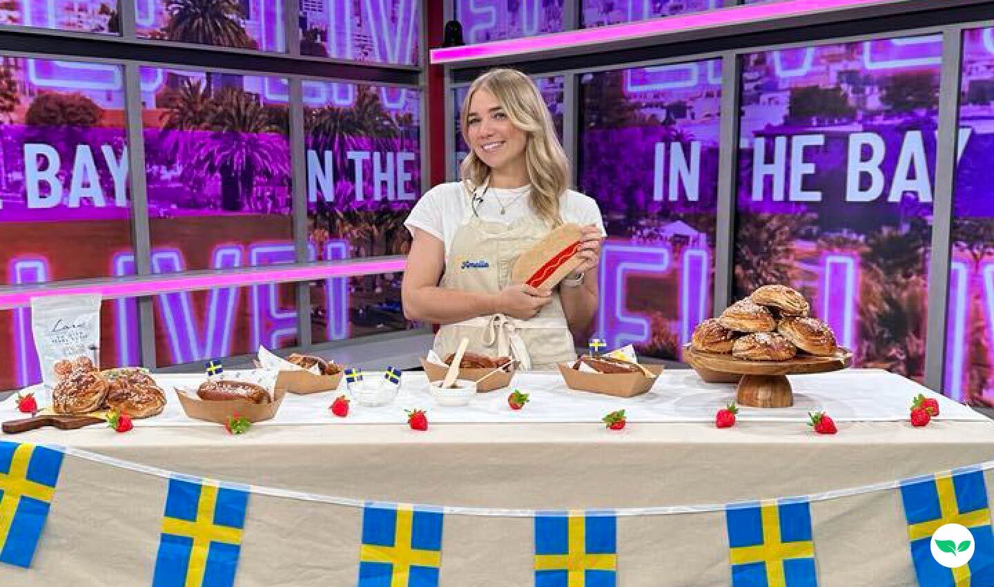 Amelia Eudailey on set of Live in the Bay, standing behind a decorated table of Swedish pastries and hot dogs.
