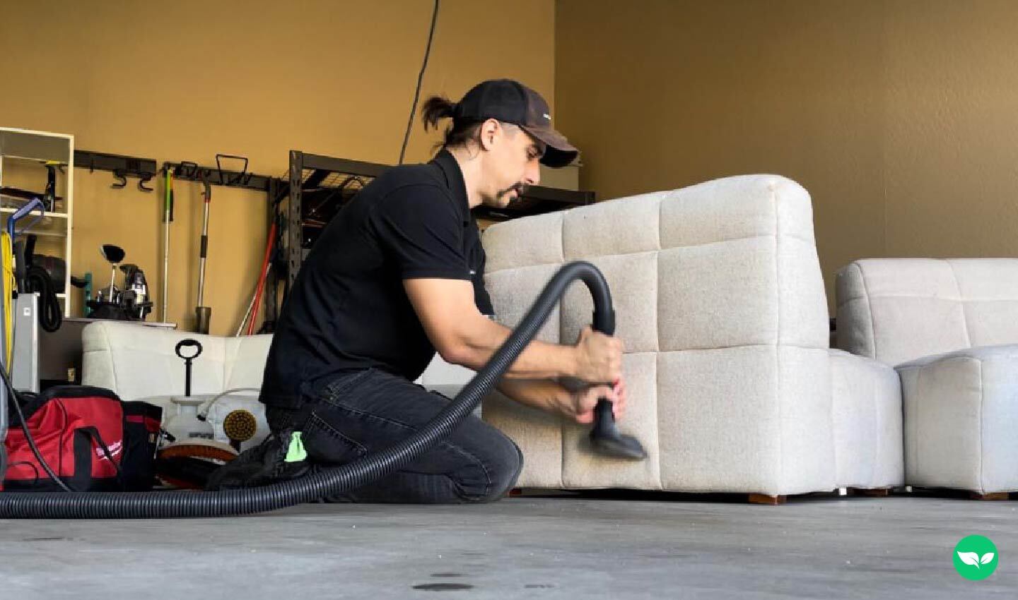 Kyle Stroud kneeling in a garage while cleaning a beige sofa with a commercial vacuum hose, surrounded by professional gear and tools.