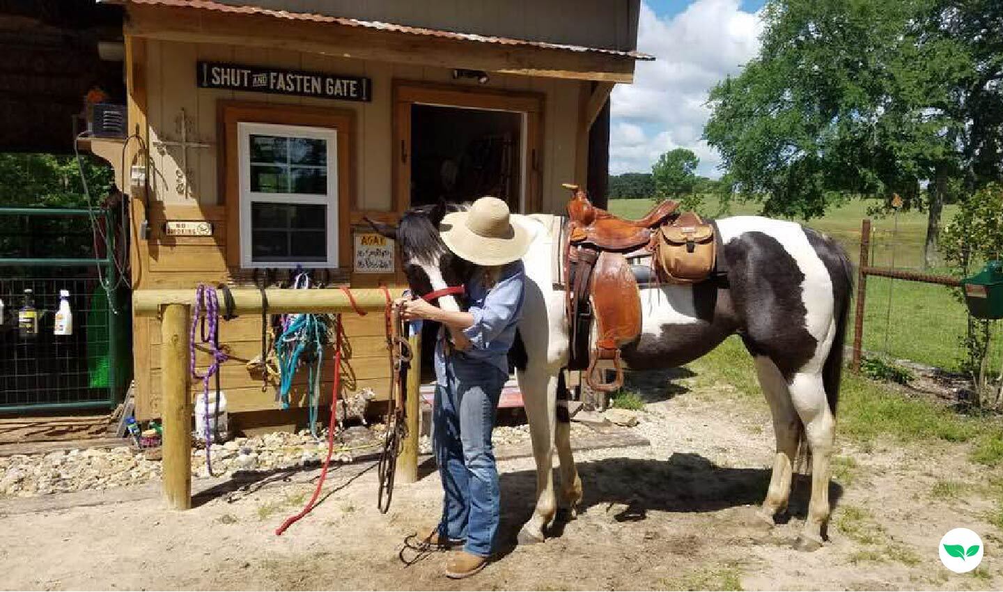 Melody Morton-Buckleair in a sun hat standing beside her saddled black-and-white horse outside a small barn.