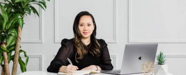 Linda Tran sits at her desk in a sunlit home office, working on a laptop with notebooks, pens, and presentation materials nearby.